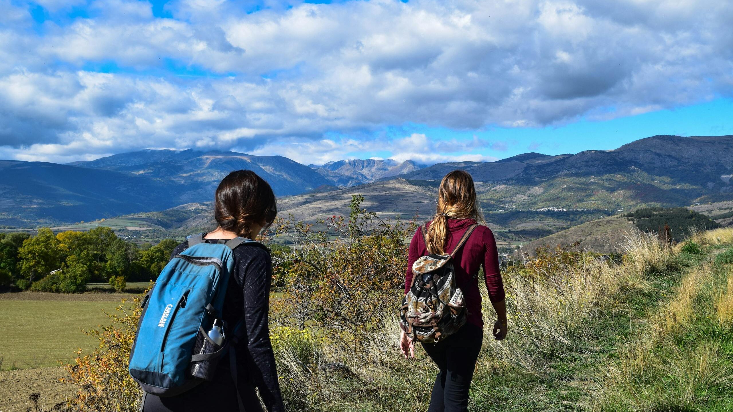 Two women hiking on a mountain trail with scenic views under a blue sky.